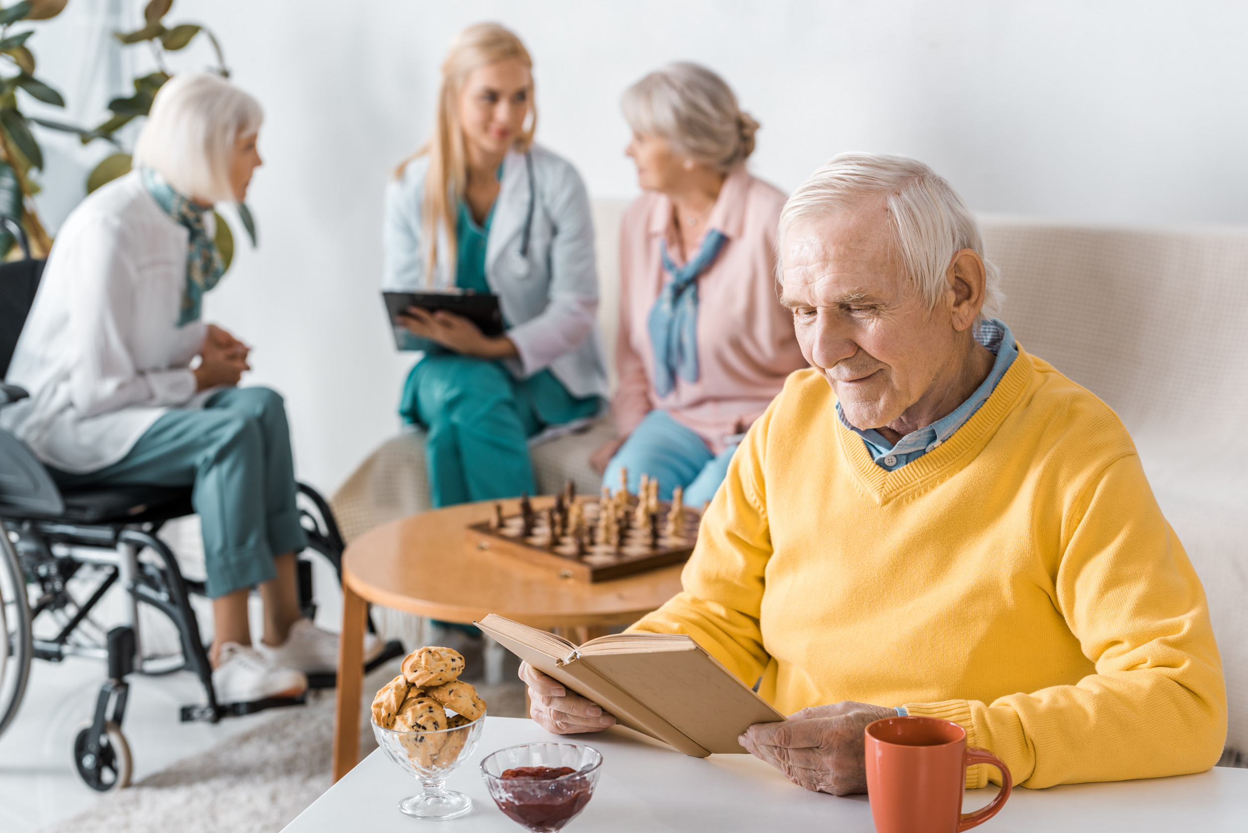 senior man reading book while female doctor examining senior women Old couple, portrait and helping man in wheelchair, home and support husband with injury